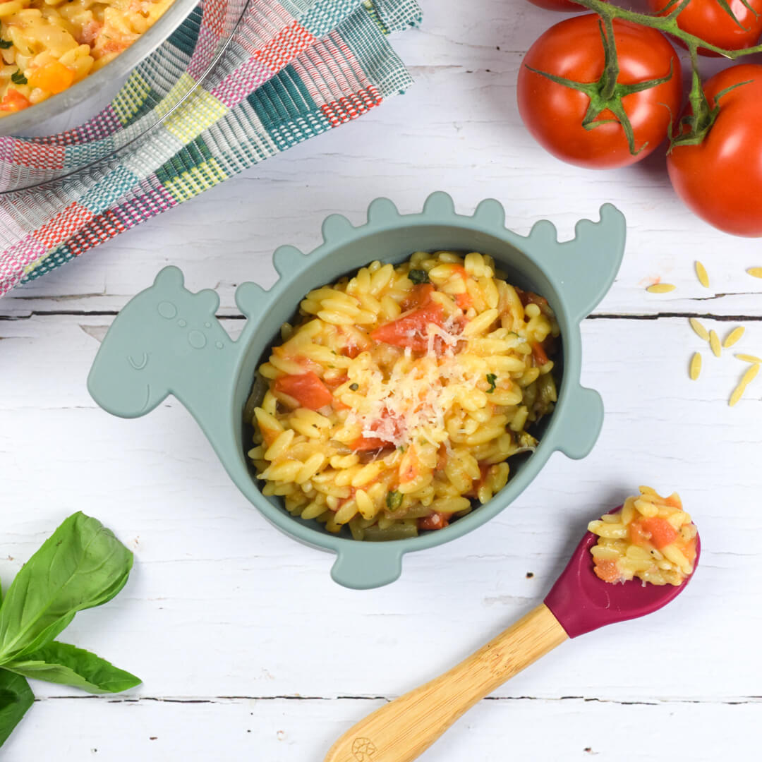 Baby dinosaur bowl with tomato orzo and a spoon with a little of the food. Also tomoatoes and large family bowl.