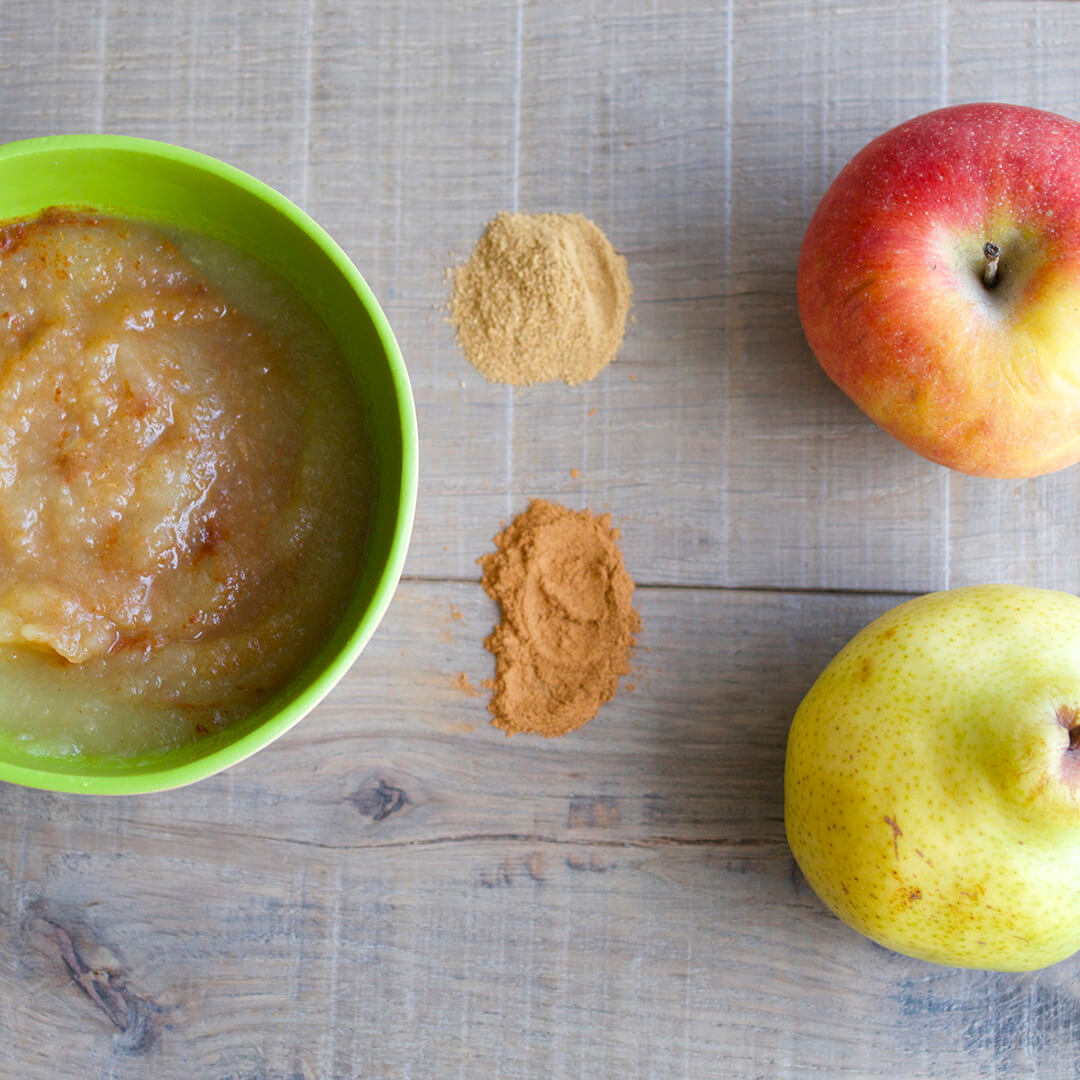 An image of apple puree in a bowl. There is an apple and a lemon next to the bowl and some spices