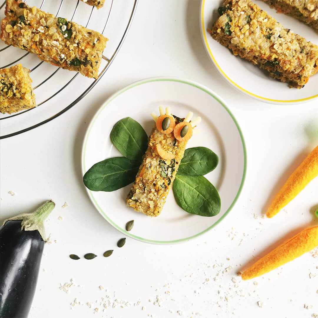 Aubergine flapjacks on plates and serving dishes. One of the aubergine flapjacks has carrot slice and pumpkin seed eyes and spinach leaves for wings with the pumpkin seed tail creating a dragonfly