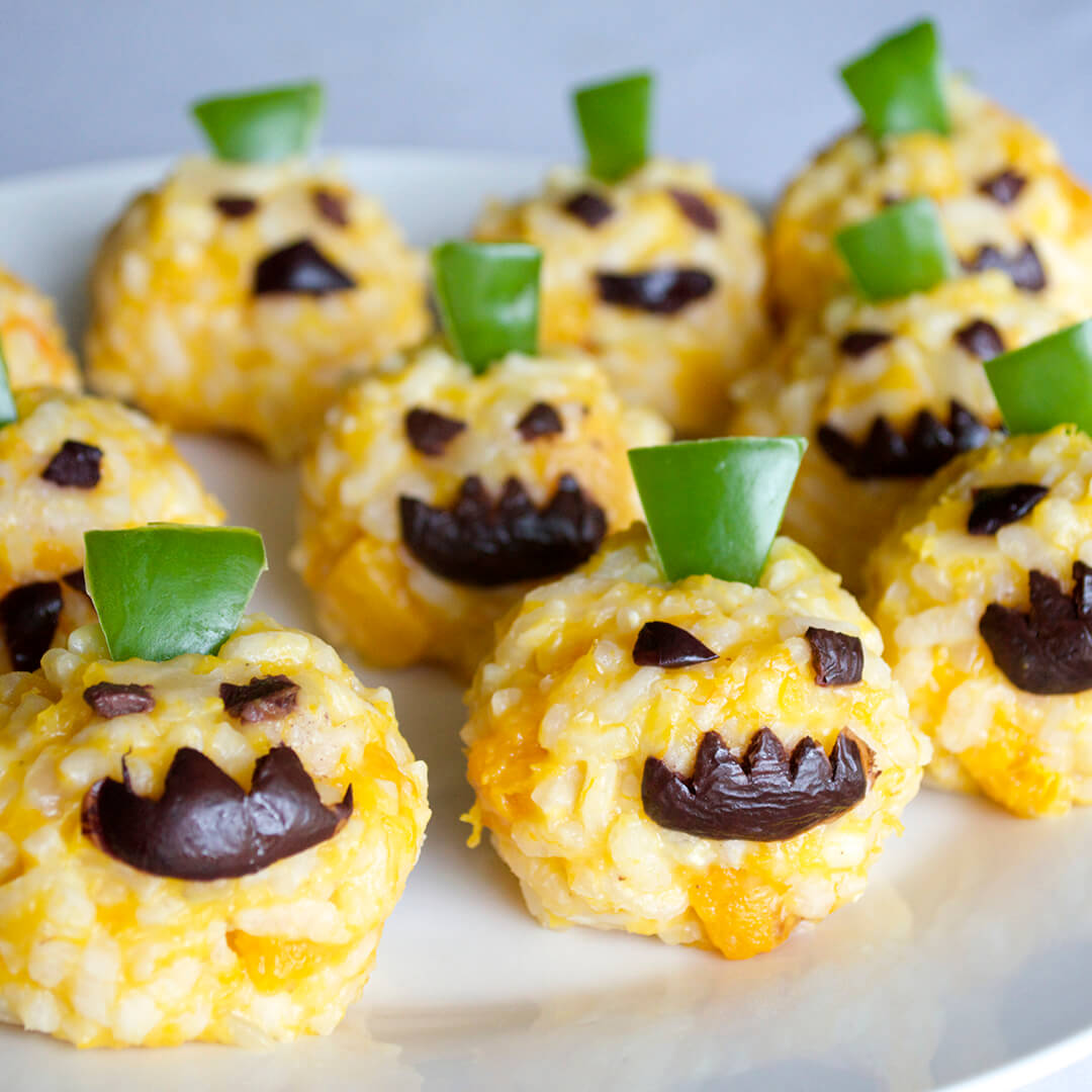 Spooky pumpkin risotto balls eyes and mouths made out of olives and a rectangle of pepper on top of their heads designed to look like pumpkins
