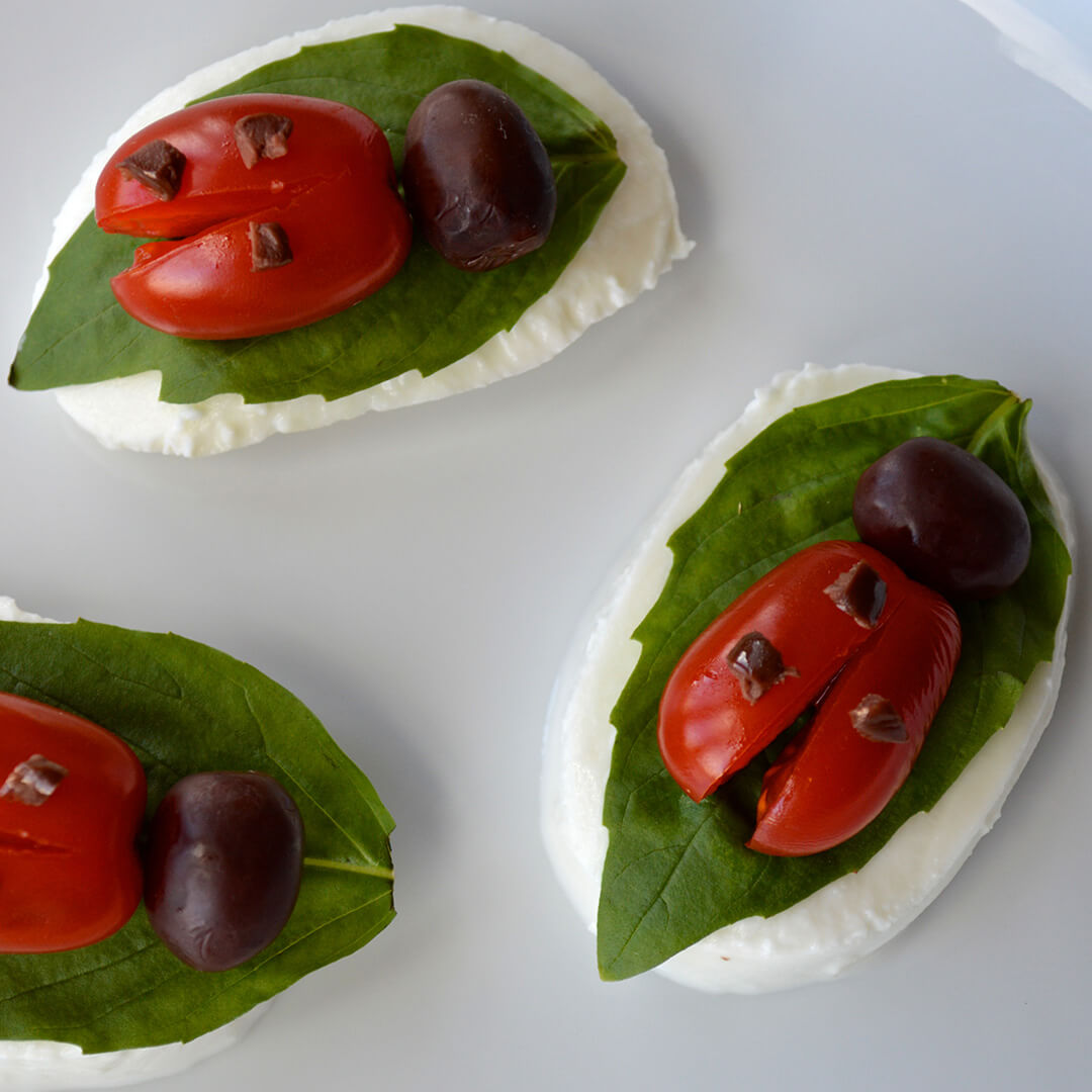 Mozzarella slices topped with basil leaves and tomatoes cut to look like ladybird wings with olive heads and olive spots