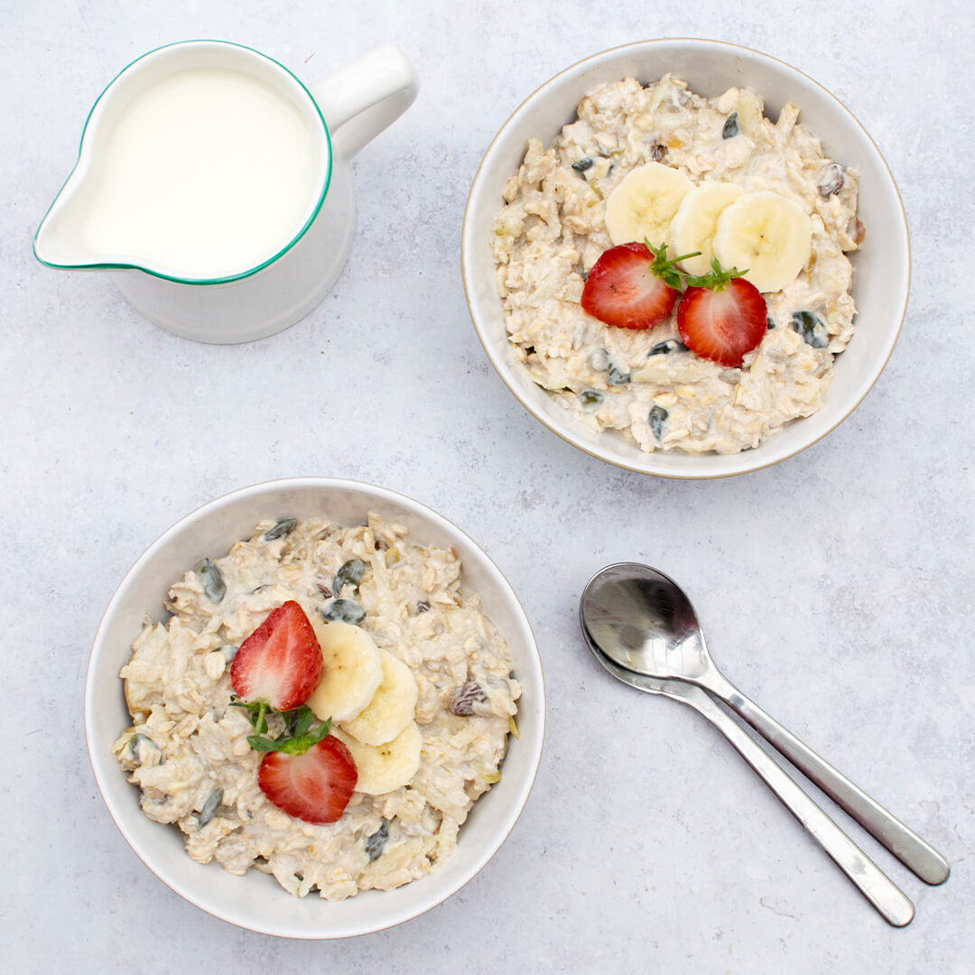 Two bowls of overnight oats topped with sliced banana and strawberries, next to a small jar of milk