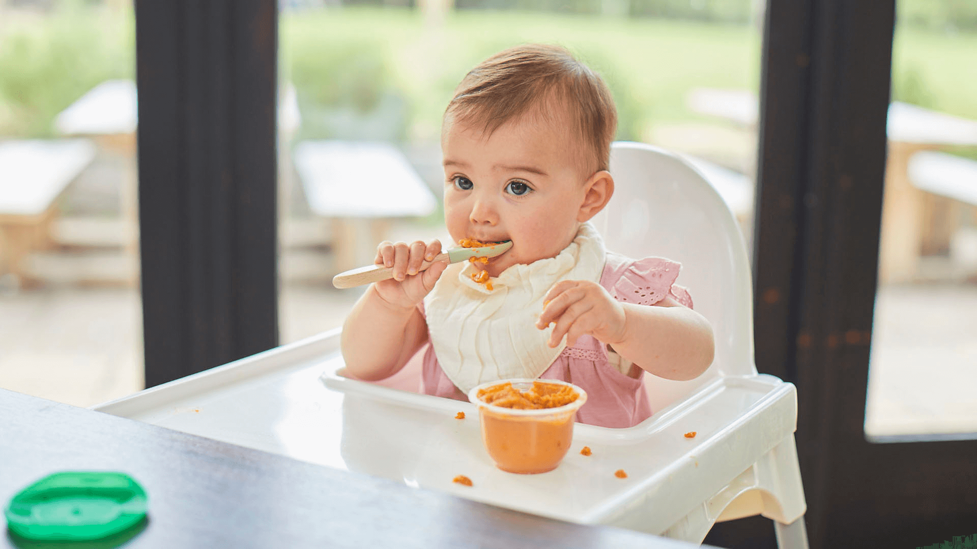 Baby on a highchair with a bib on feeding themselves from an Organix meal pot