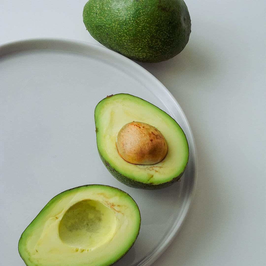 A white plate with a halved avocado next to a whole avocado