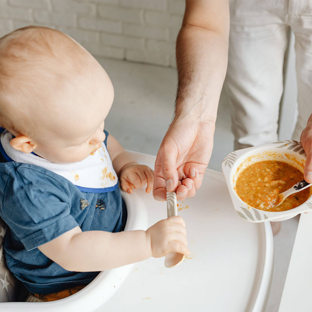 A baby on a high chair being handed a spoon and a bowl of food