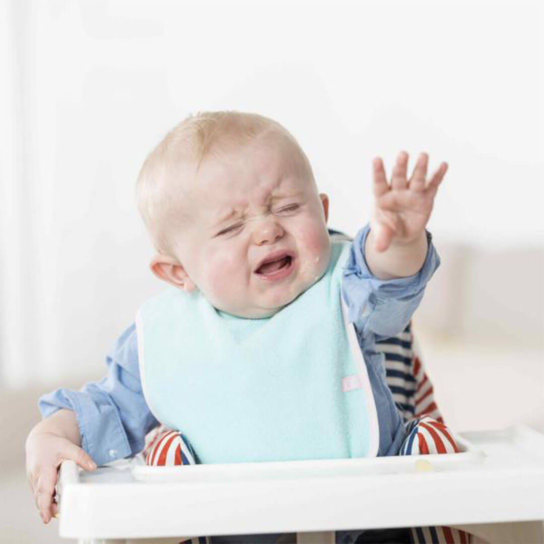 A toddler sitting on a high chair, crying with his hand stretched out