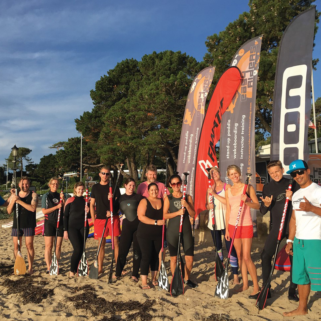 12 people stood on the beach holding up paddles stood in front of paddle boars and tall banners. They are all wearing beach wear and wetsuits with sunglasses on. There are trees in the background and a blue sky