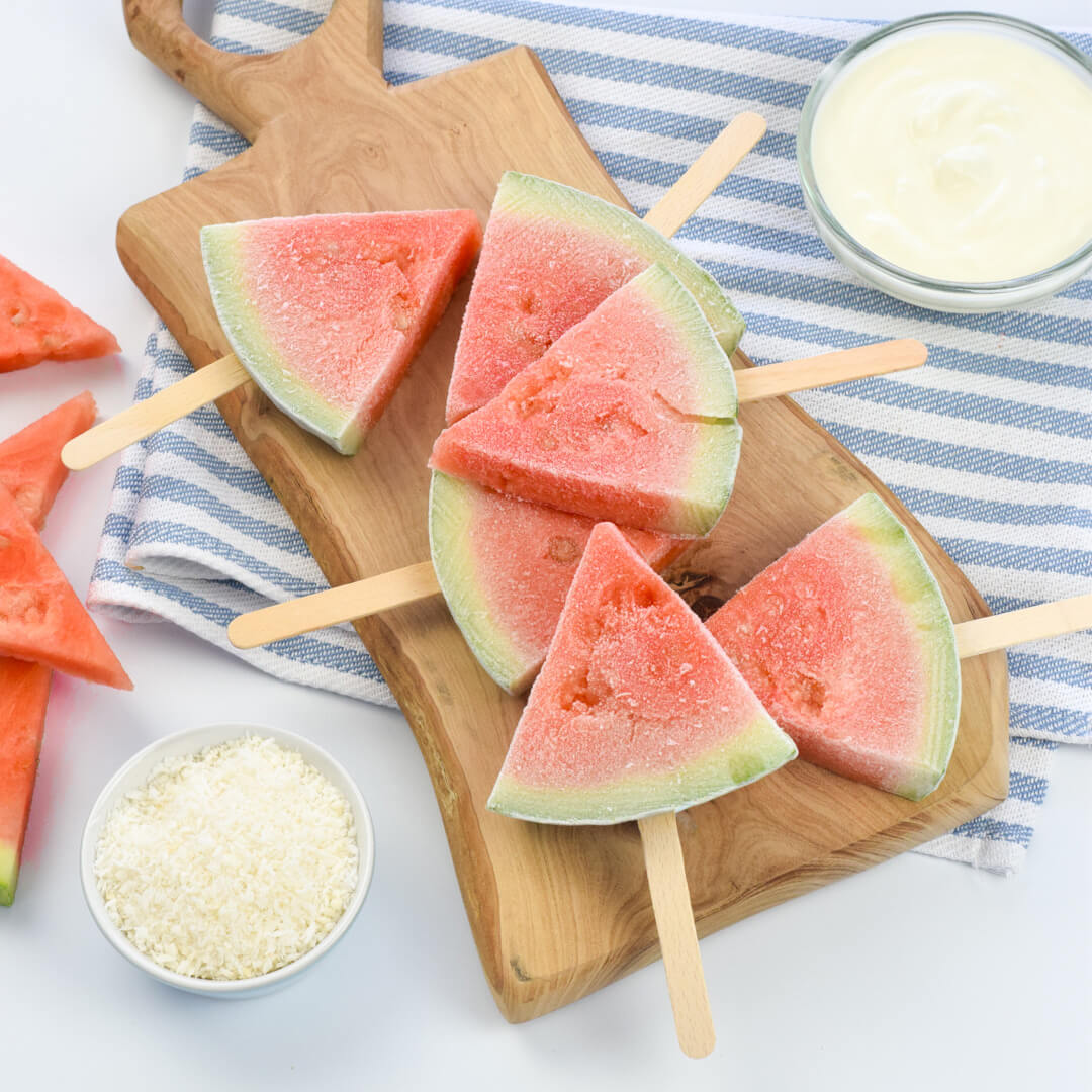 A chopping board with 6 frozen watermelon pops served with a bowl of natural yoghurt and a bowl of desiccated coconut 