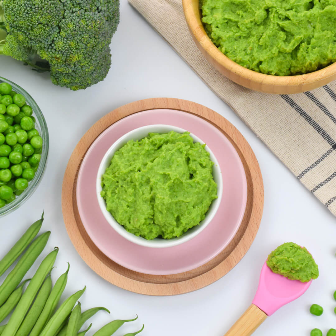 A serving of green bean, pea and broccoli mash next to a bowl of green bean mash, some peas, green beans and a broccoli floret