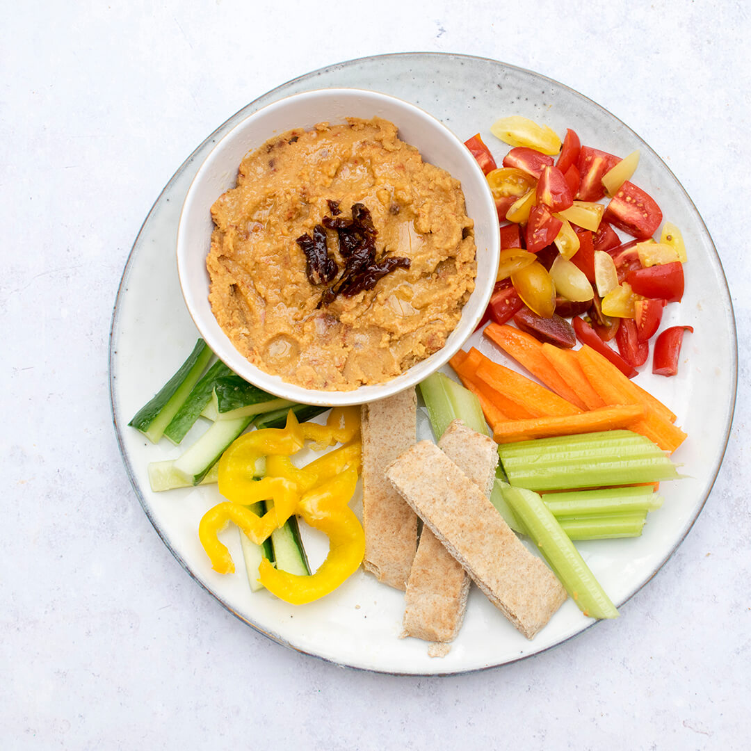 A small bowl of red lentil dip served with flatbread, carrot and cucumber sticks, peppers and tomatoes