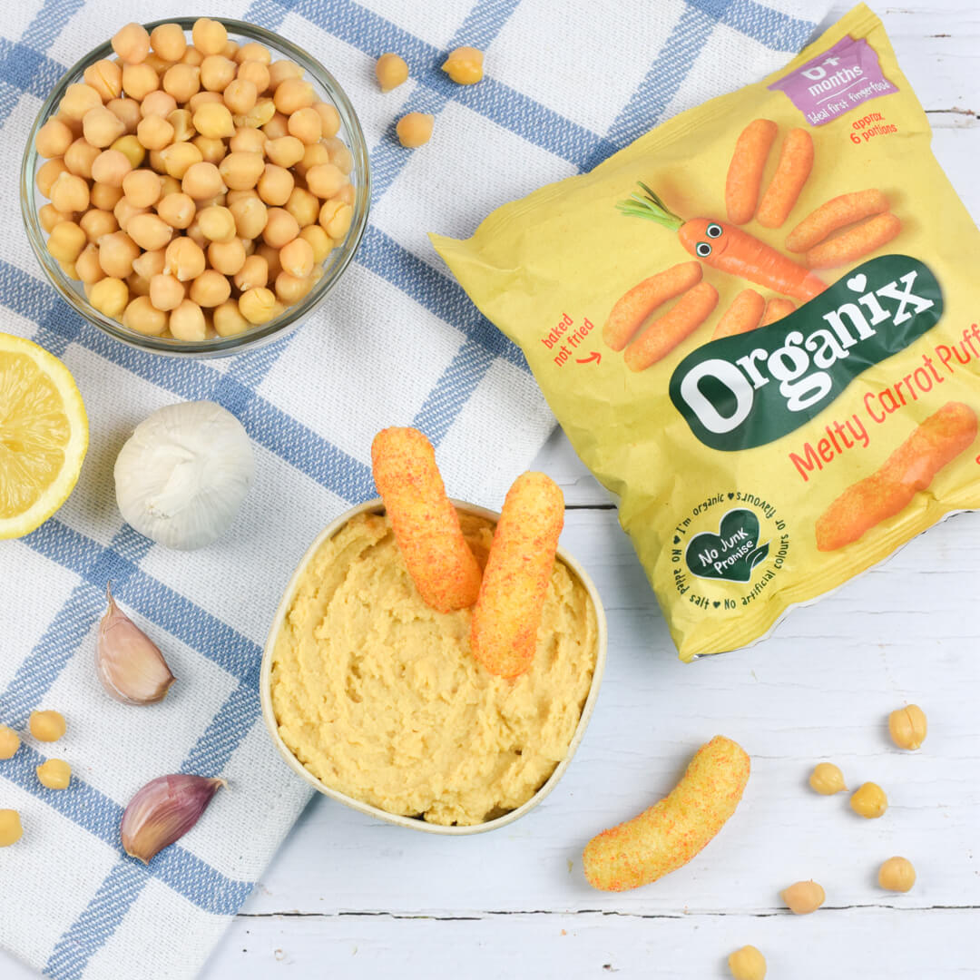 A bowl of hummus next to a bowl of chickpeas, some garlic cloves, a halved lemon and some Organix melty carrot puffs