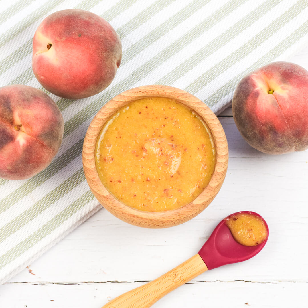 A bowl of peach puree next to 3 whole peaches