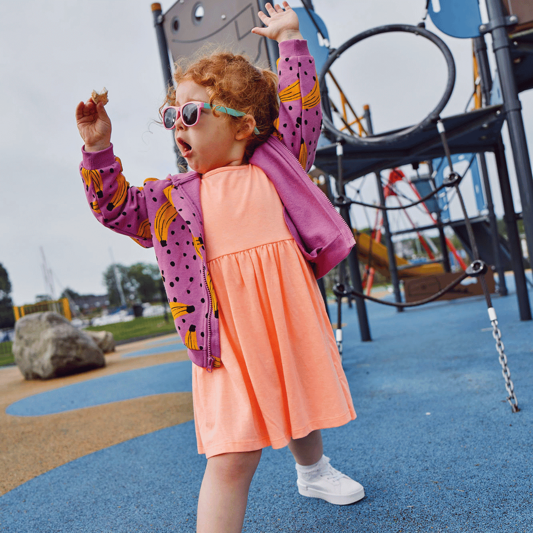 Little girl dressed in colourful clothes, an orange dress and a pink jacket with black spots. She's also wearing sunglasses. She's at a play area and has her hands up in the air in excitement