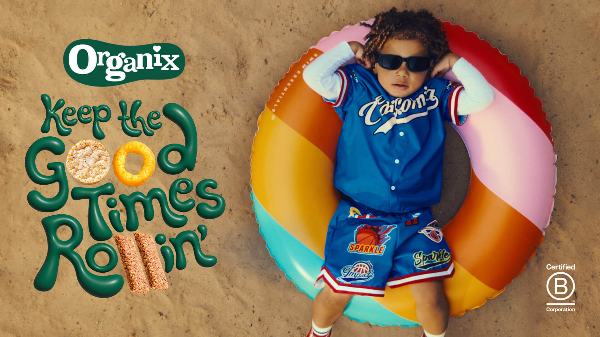 A toddler in a sandy beach laying down on a ring inflatable with their hand on their head. The child looks 'cool' and is wearing sunglasses. 