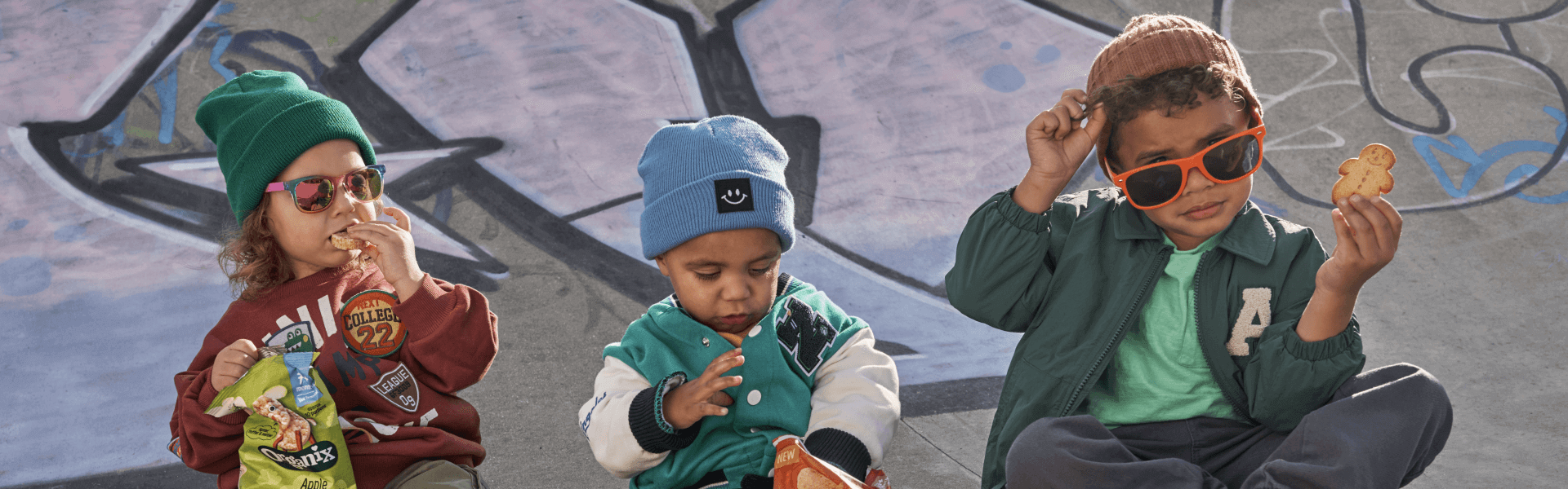 Three toddlers sittng outdoors at a skate park eating Organix Snacks, dressed in urban clothes