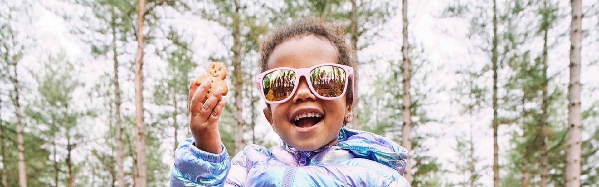 Little child holding an Organix Banana Bread Biscuit in their hand. They are outdoors, in a woodland area, and is wearing winter clothes and sunglasses. 