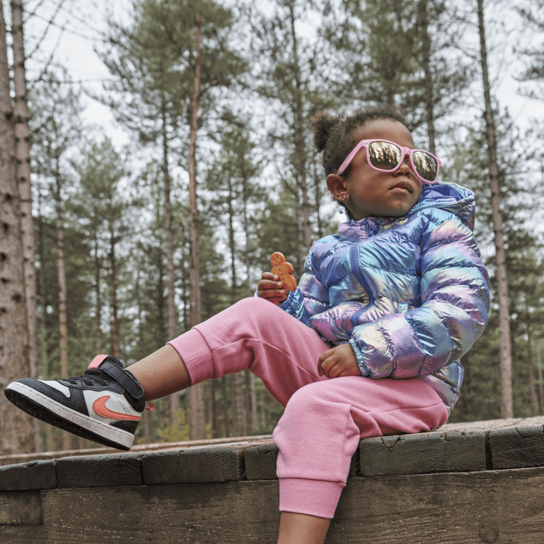 A child sitting on a bench outdoors, wearing cool winter clothes and sunglasses, and holding an Organix biscuit