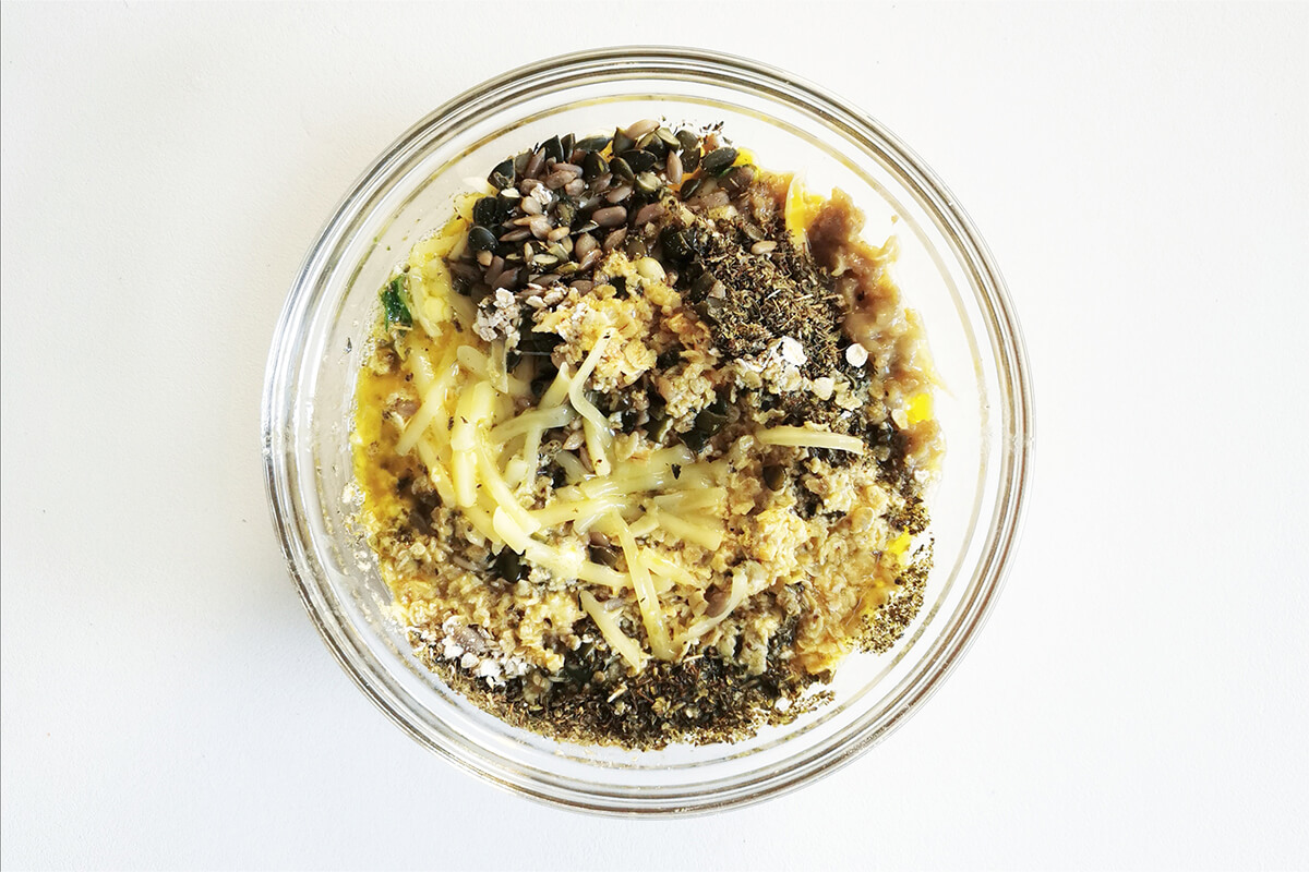 A glass bowl with grated carrot, grated aubergine, chopped spinach, grated cheese, oats, eggs, melted butter, herbs and seeds