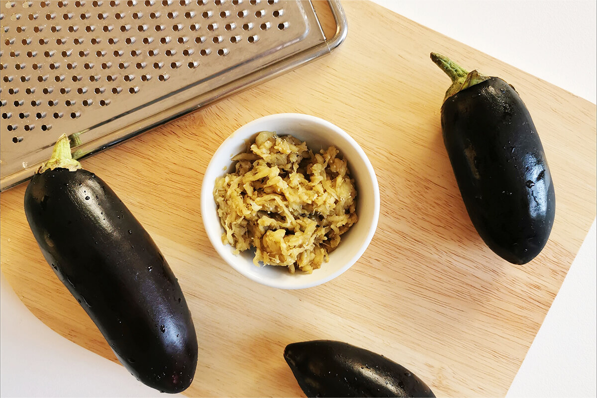 A dish of grated aubergine on a chopping board next to a metal grater and 3 aubergines