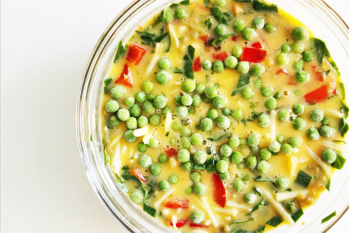 A glass bowl of beaten eggs combined with grated aubergine, chopped peppers, spinach, frozen peas, baby tomatoes and grated cheese