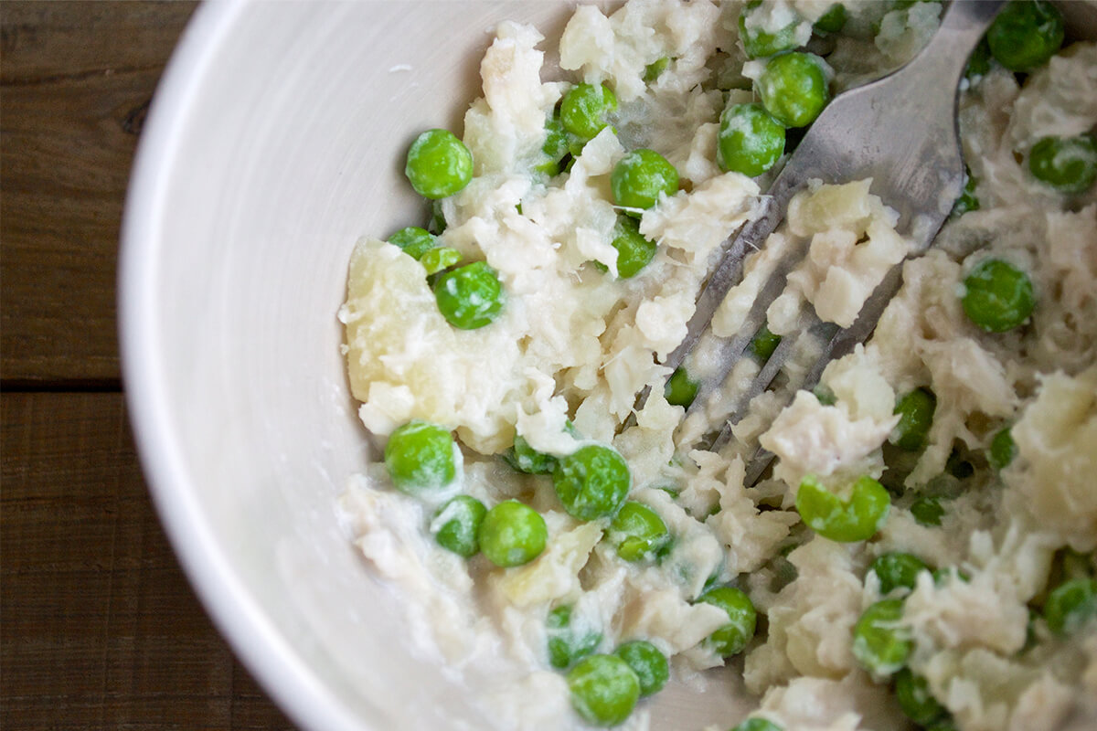 A bowl of fish, potatoes and peas being mashed with a fork