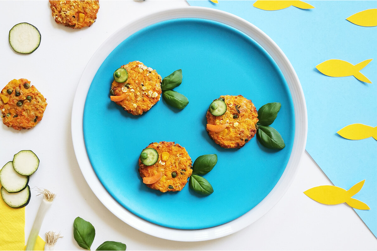 3 Baby Salmon Fish Cakes on a plate. The fish cakes have cucumber and peas for eyes a carrot mouth and basil leaves for tails