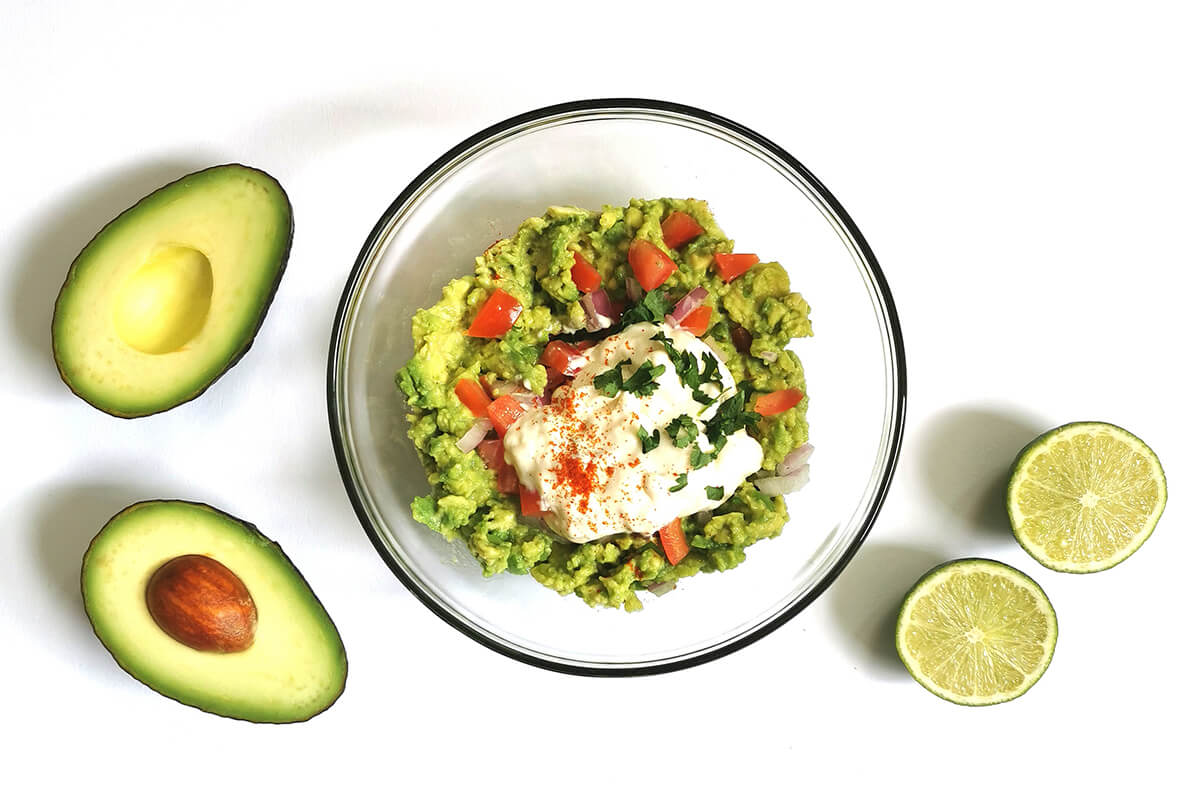 A glass bowl with smashed avocado, chopped red onion and tomatoes, Greek yoghurt, smoked paprika and coriander, next to a halved avocado and halved lime
