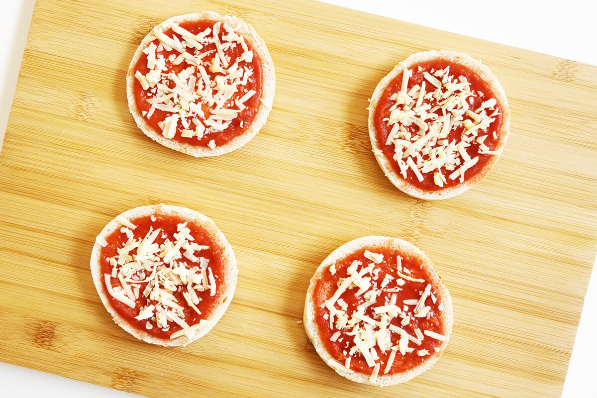 2 halved muffins on a chopping board, topped with pizza sauce and cheese