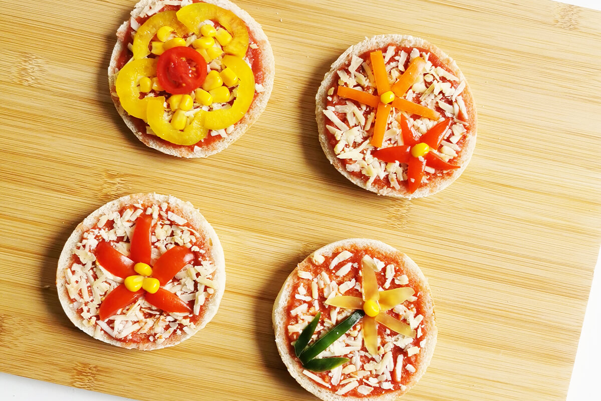2 halved muffins on a chopping board, topped with pizza sauce and cheese and some flower shapes made out of vegetables