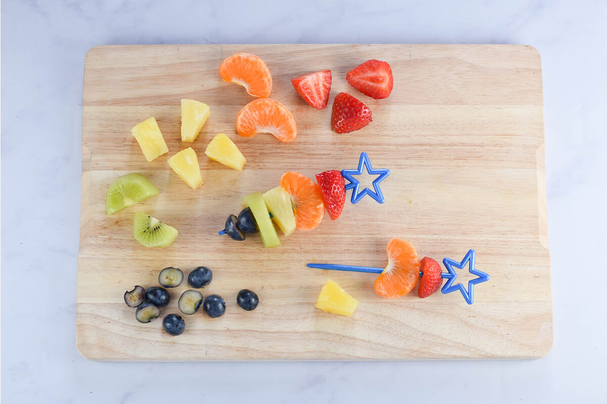 Strawberries, blueberries, kiwi, clementine and pineapple chunks on a chopping board being put onto skewers