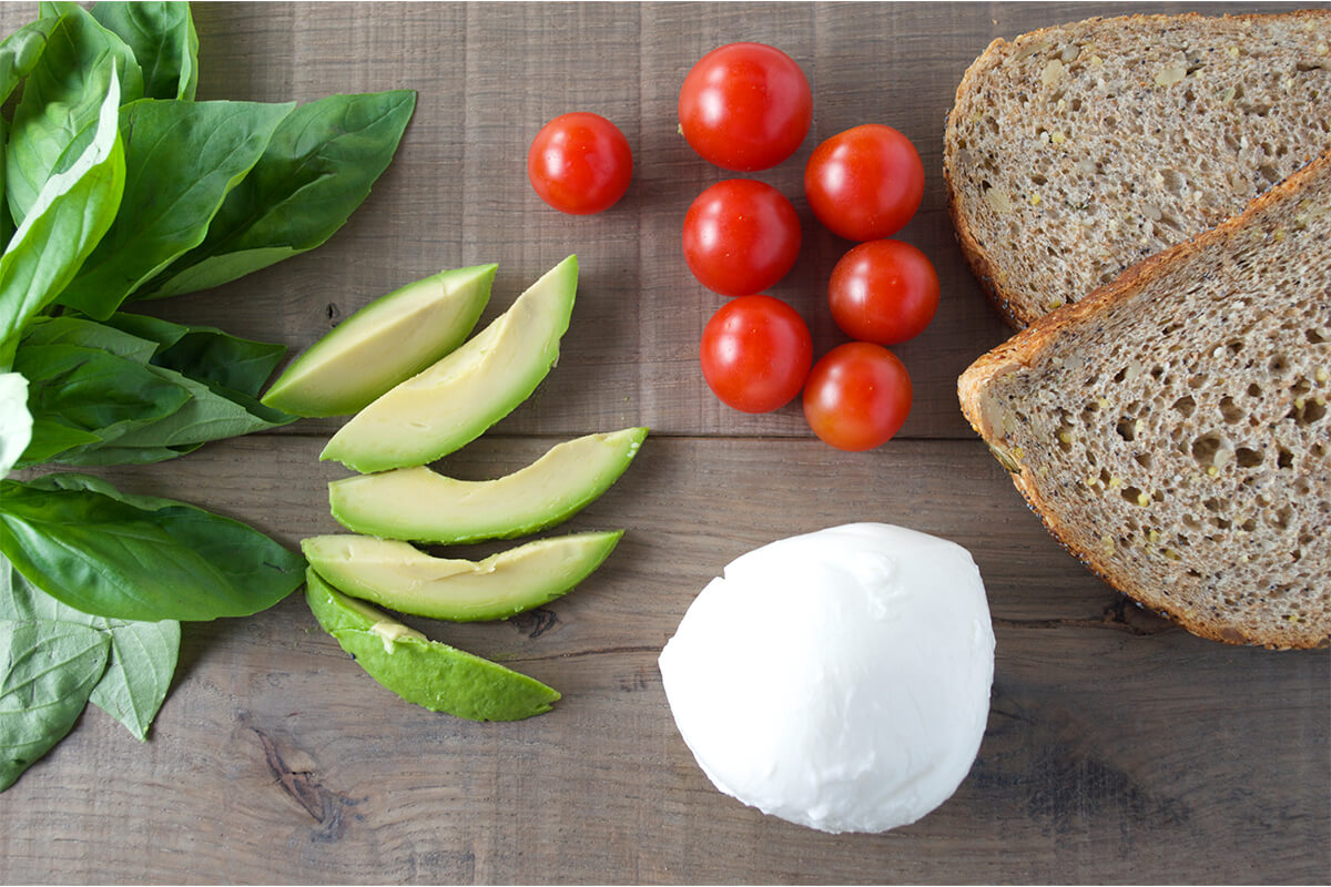 Basil leaves, sliced avocado, cherry tomatoes, a ball of mozzarella sand 2 slices of bread on a table