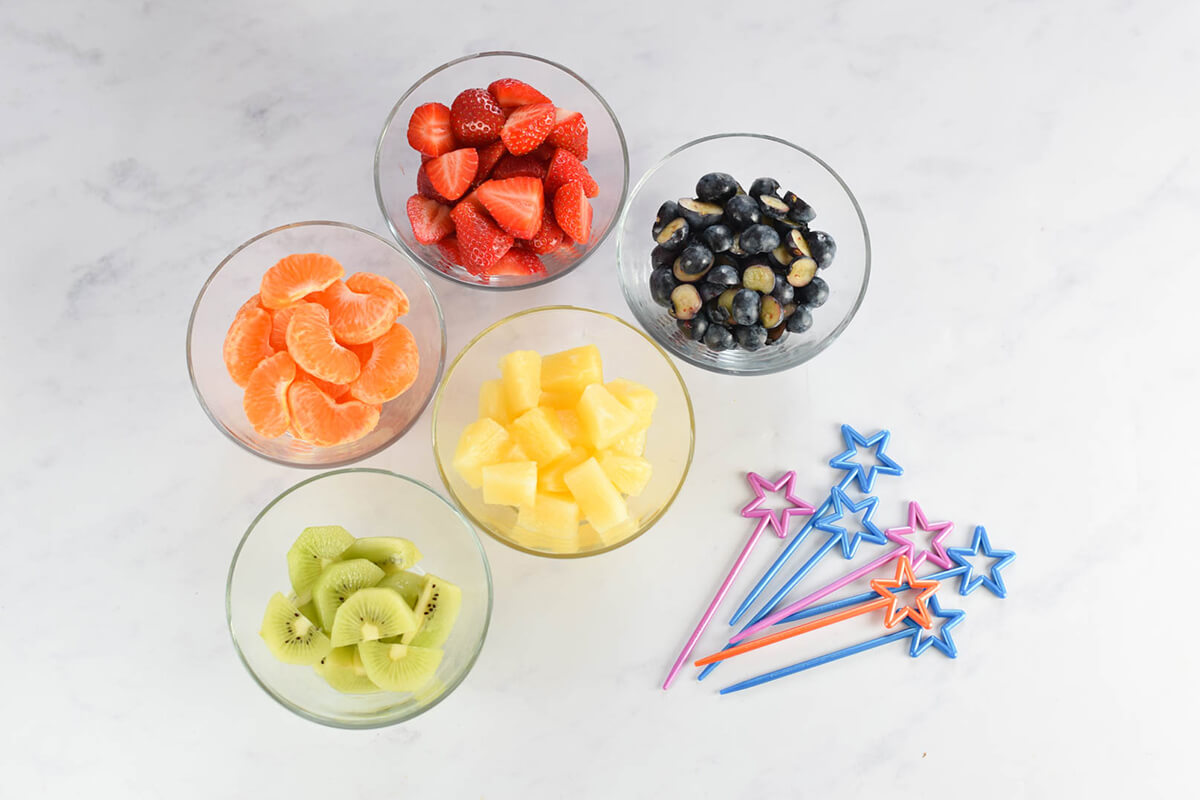 Small glass bowls of strawberries, clementines, pineapple chunks, kiwi and blueberries next to some skewers