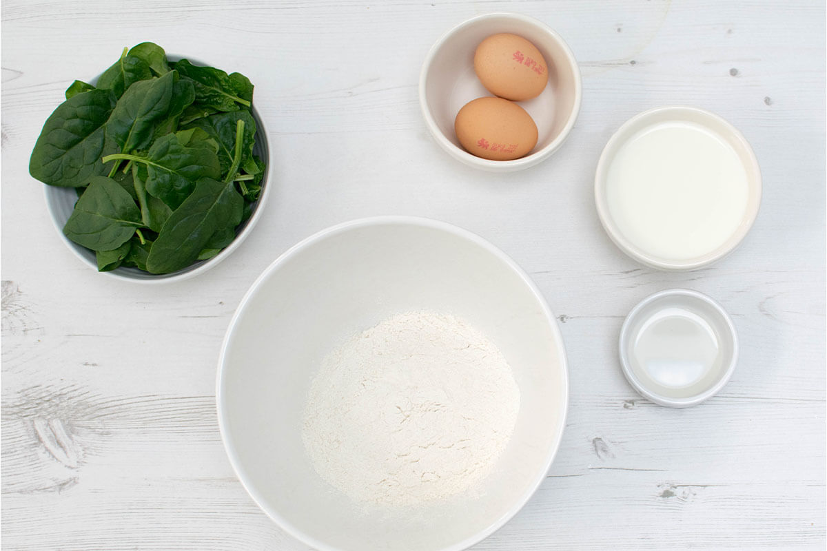 A bowl of spinach next to bowls of eggs, milk, rapeseed oil, and flour