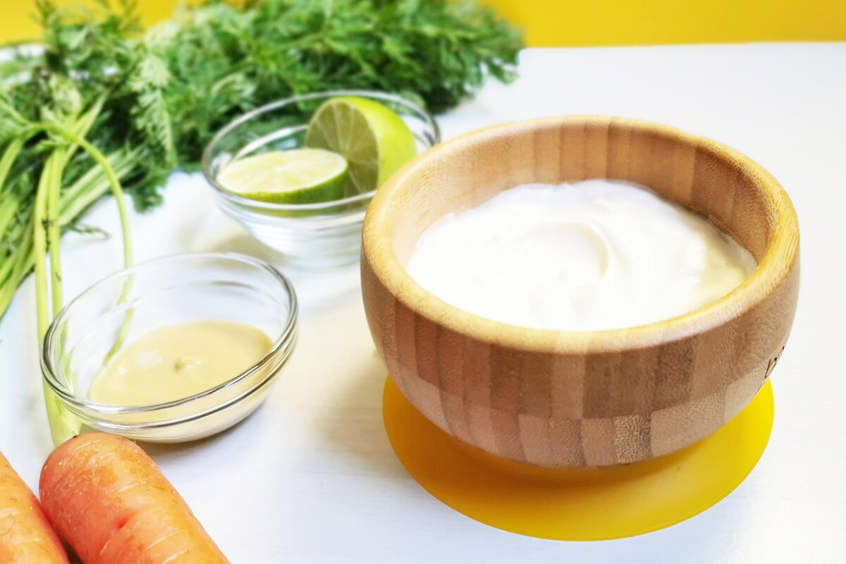 A bowl of crème fraiche next to small bowls of mustard and halved limes, next to some carrots