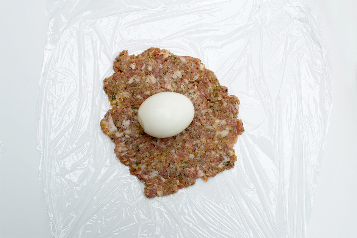 A square of cling film with meat mix rolled out onto the middle of it, topped with an egg