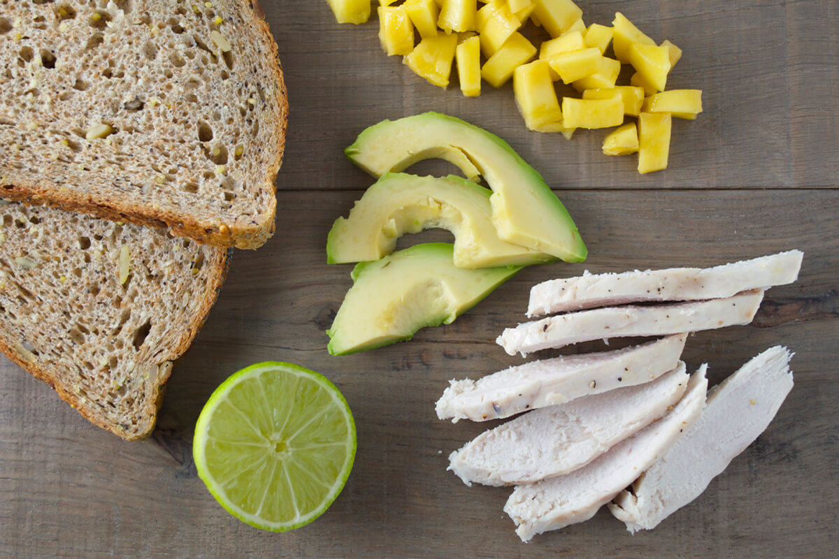 A chopping board with 2 slices of bread, mango chunks, sliced avocado, chicken strips and a halved lime