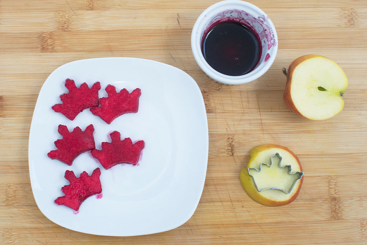 Crown shaped apple slices being cut out with a crown shaped cutter. 5 of them have been soaked in beetroot juice to give them a red colour