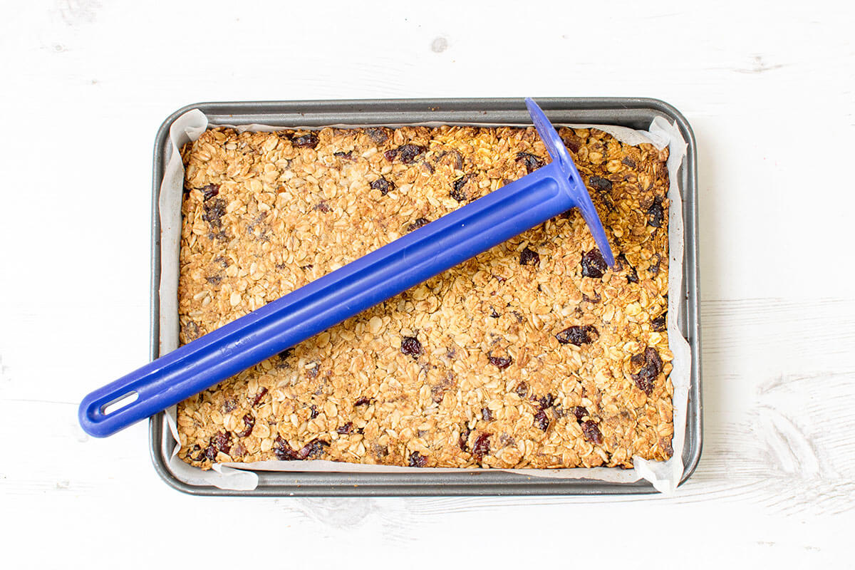Cooked date & cranberry seed mix in a lined baking tray with a blue potato masher on top of it