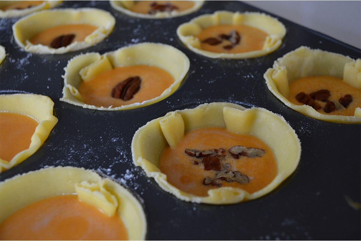 A cupcake tin with pastry filled with pumpkin mixture and topped with chopped pecans