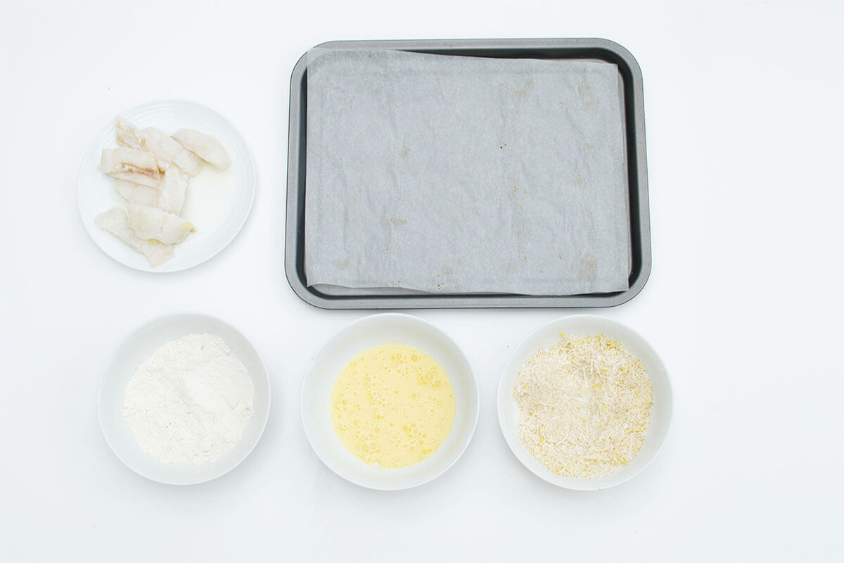 A baking tray lined with parchment paper next to a plate of fish strips, a bowl of flour, a bowl of beaten egg and a bowl of breadcrumbs mixed with cheese and lemon zest