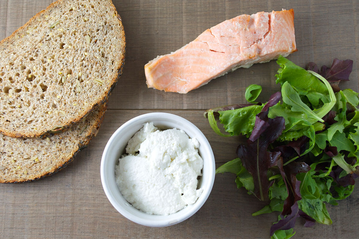 A table with bread, a salmon fillet, small ramekin of cream cheese and salad leaves