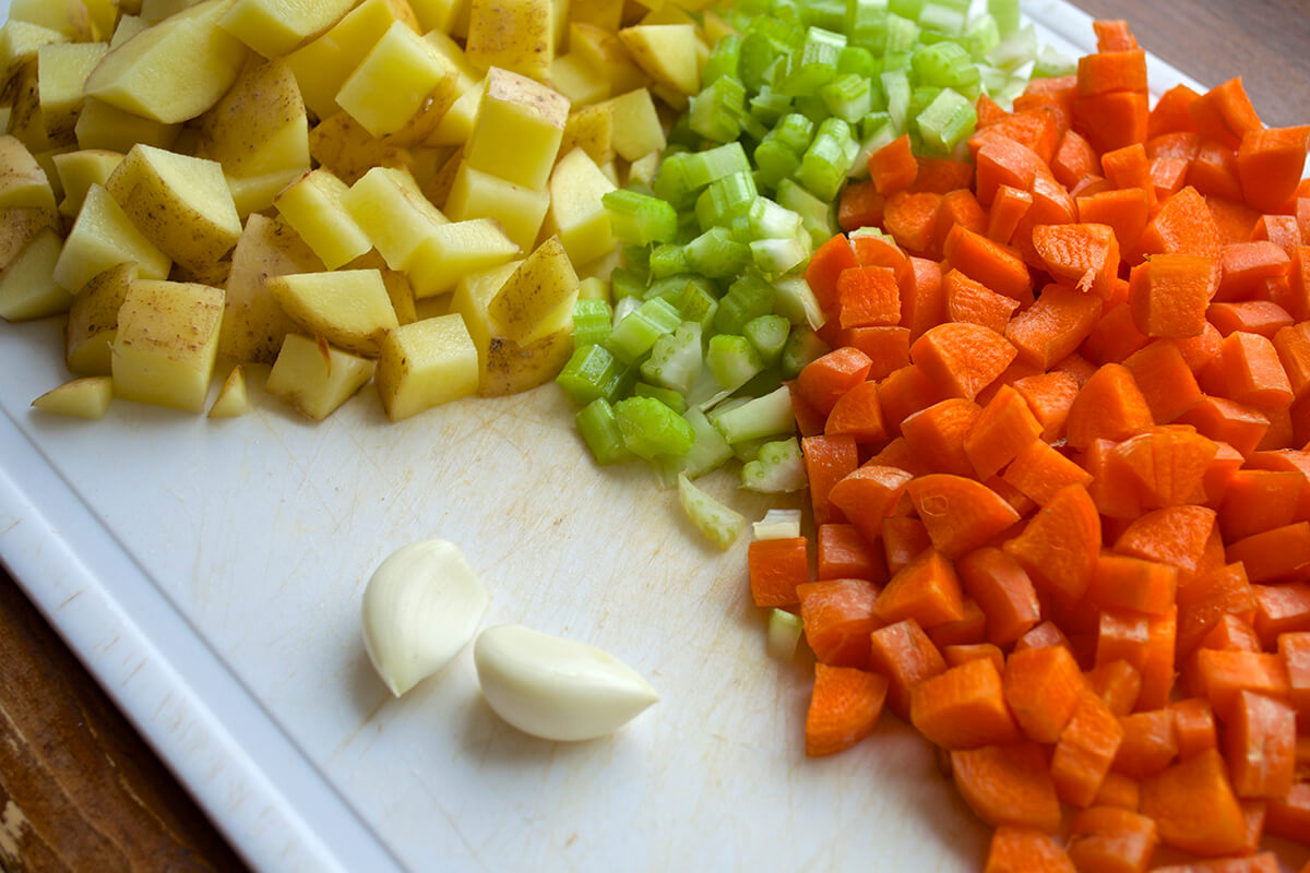 A chopping board with chopped potatoes, carrots, celery and 2 cloves of garlic