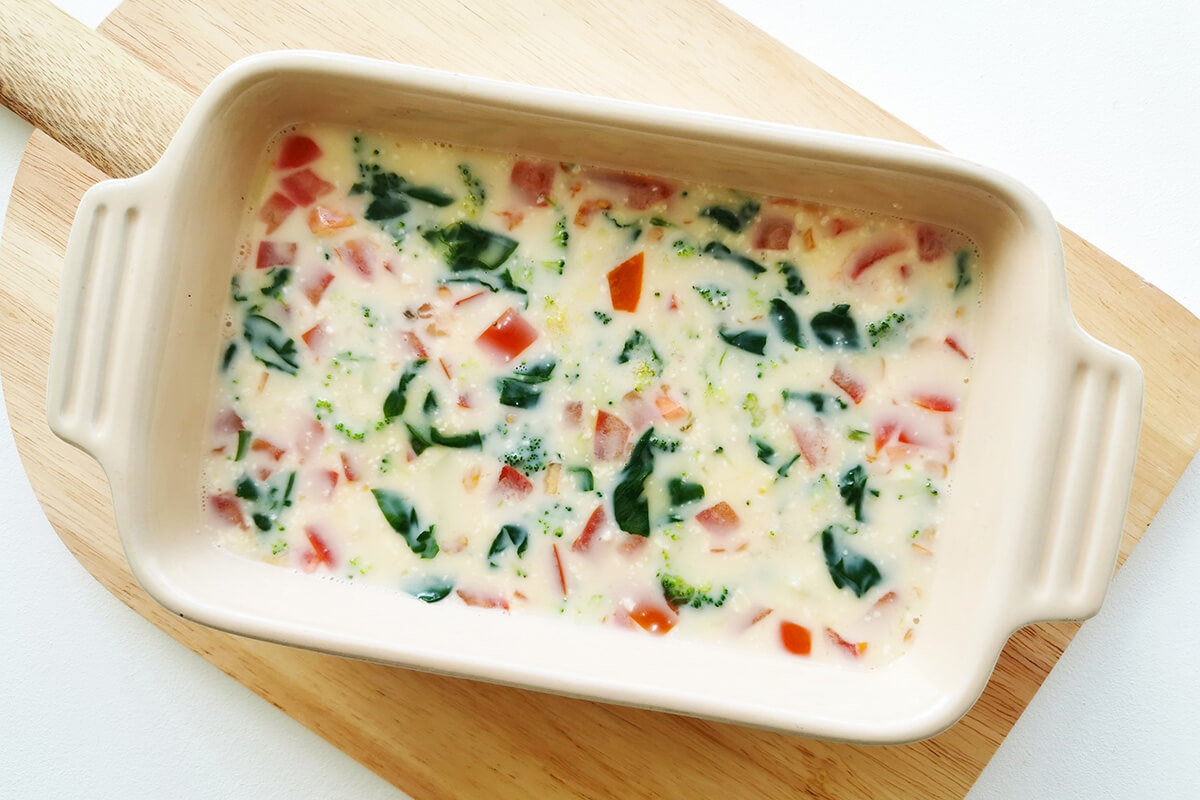 Whisked eggs and milk in a baking dish with broccoli, spinach, tomatoes and grated cheese