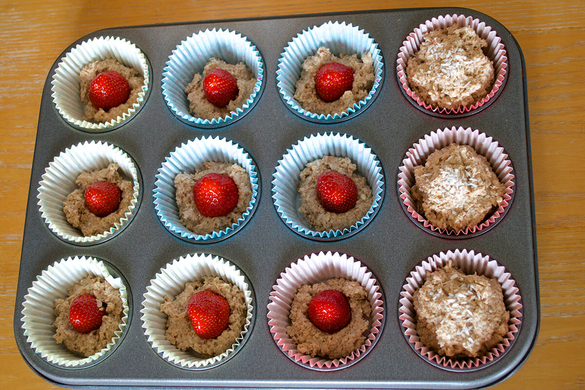 A muffin tin with paper cake cases with a spoon of muffin batter. 9 of the muffin cases have strawberries on top and haven't been layered with more muffin mix. 3 of the muffins have been covered with more muffin mix and sprinkled with desiccated coconut