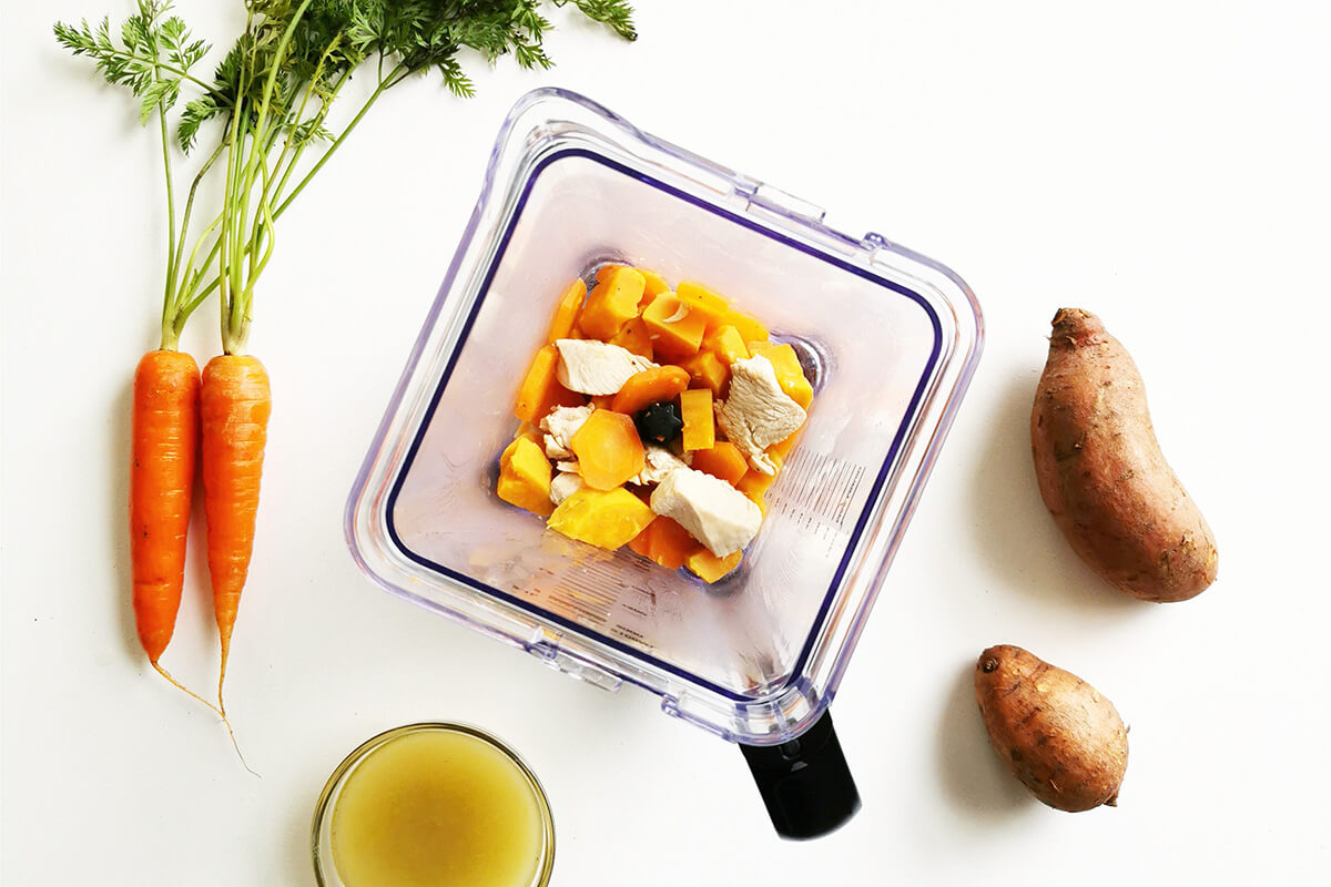 Cooked sweet potato, carrot and chicken in a blender next to some raw carrots, sweet potato and vegetable stock