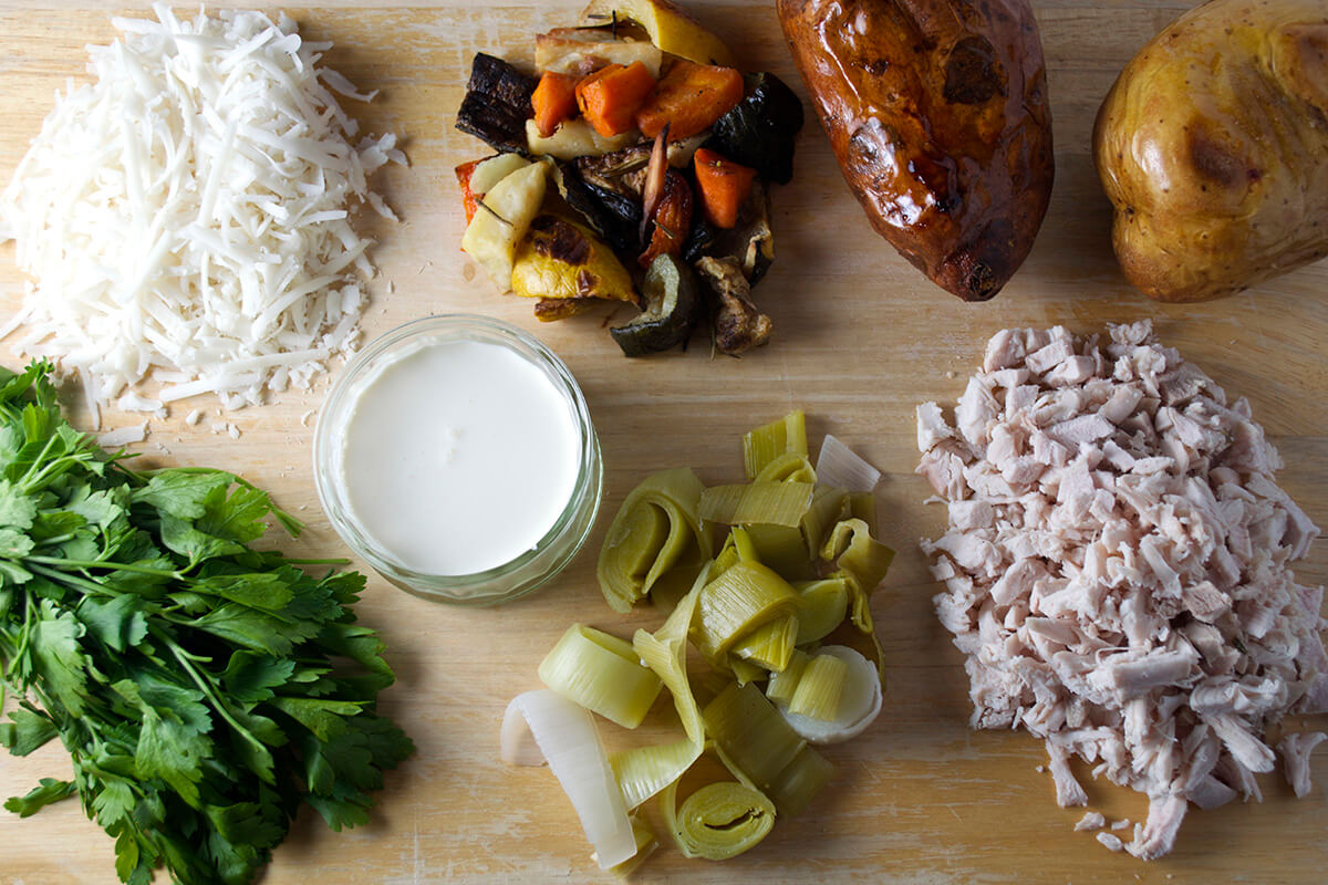 A chopping board with a baked jacket and sweet potato, vegetables, chicken/turkey, chopped leeks, parsley, cream and cheese