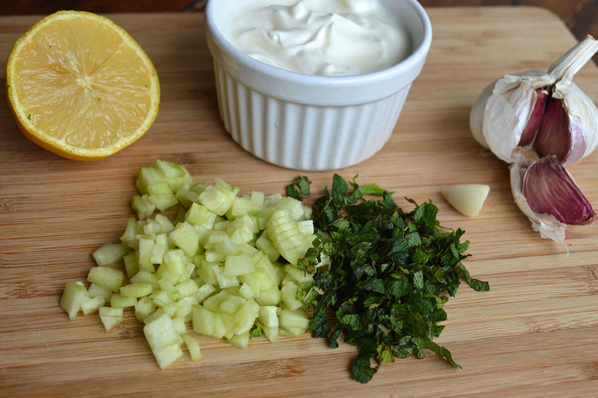A chopping board with a bulb of garlic, yoghurt, half a lemon, chopped mint and cucumber