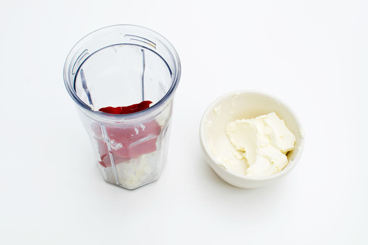 A bowl of cream cheese next to a blender with cream cheese and red pepper