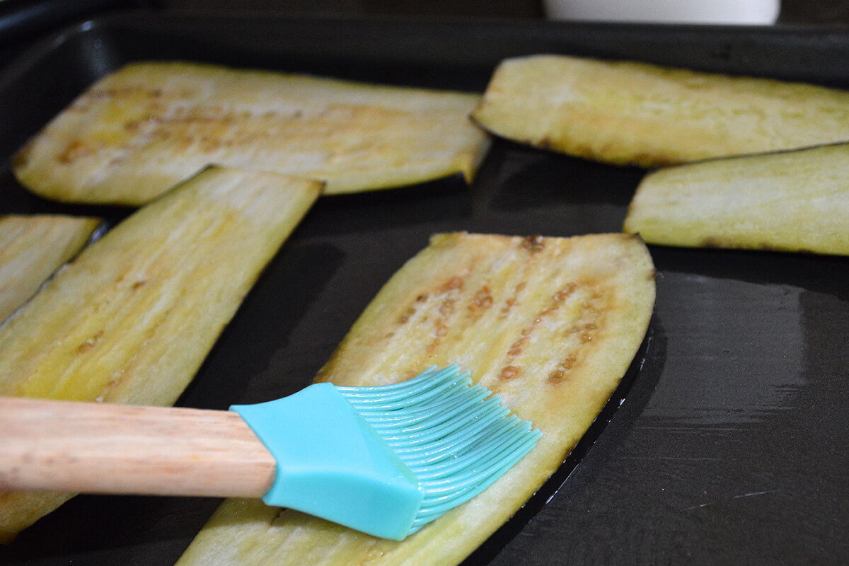 Sliced aubergine, cut lengthways, on a baking tray and being oiled with olive oil