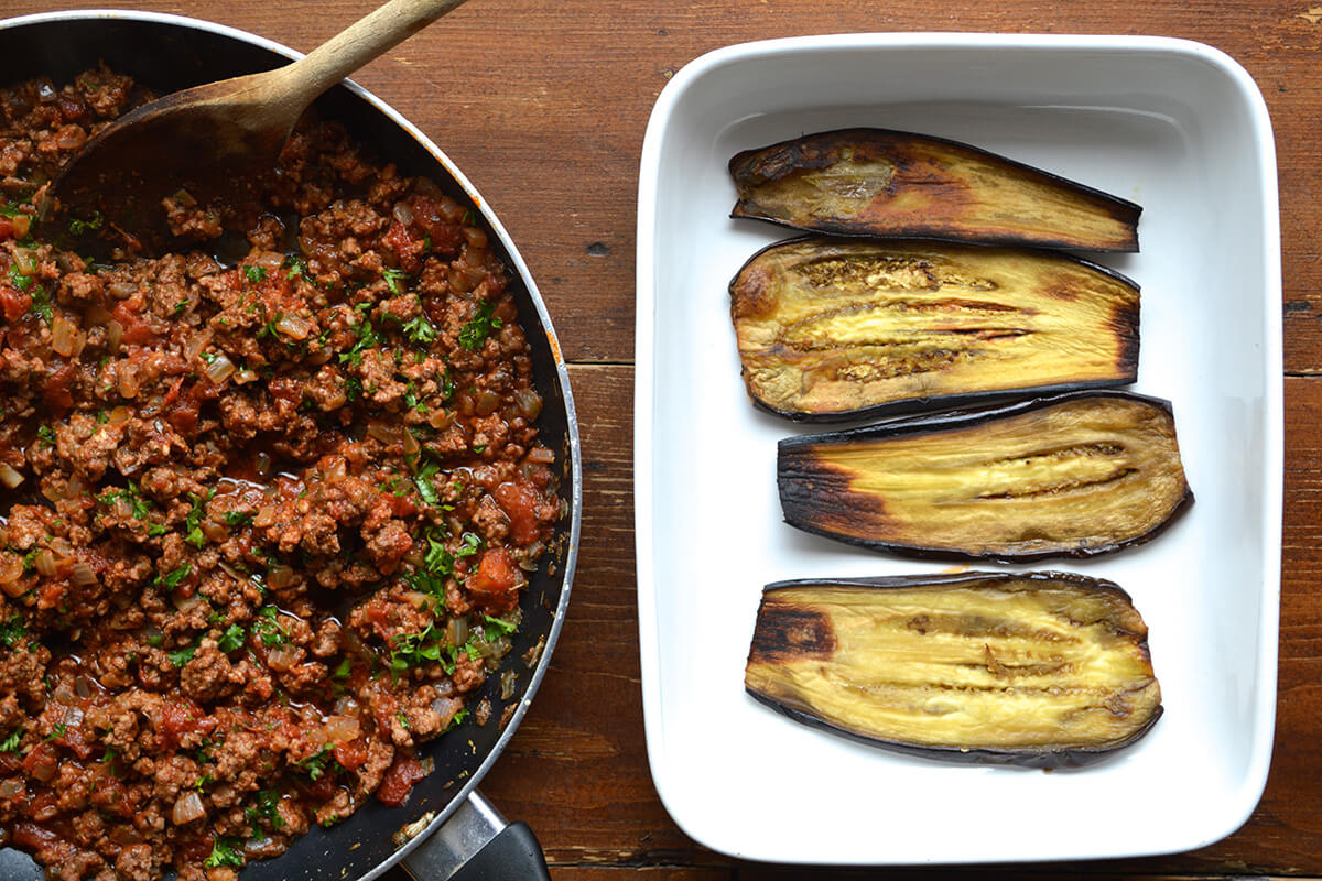 A casserole dish with four aubergine slices next to each other and a pan of fried minced lamb next to it